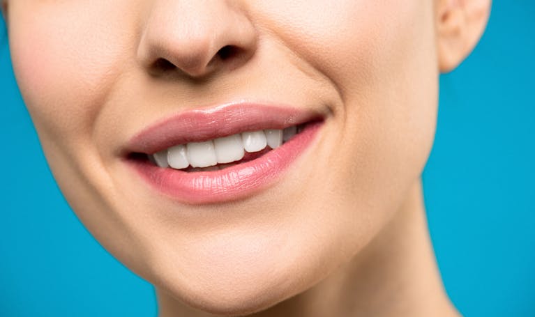 A close-up image of a woman's smile with pink lipstick and white teeth against a blue background.